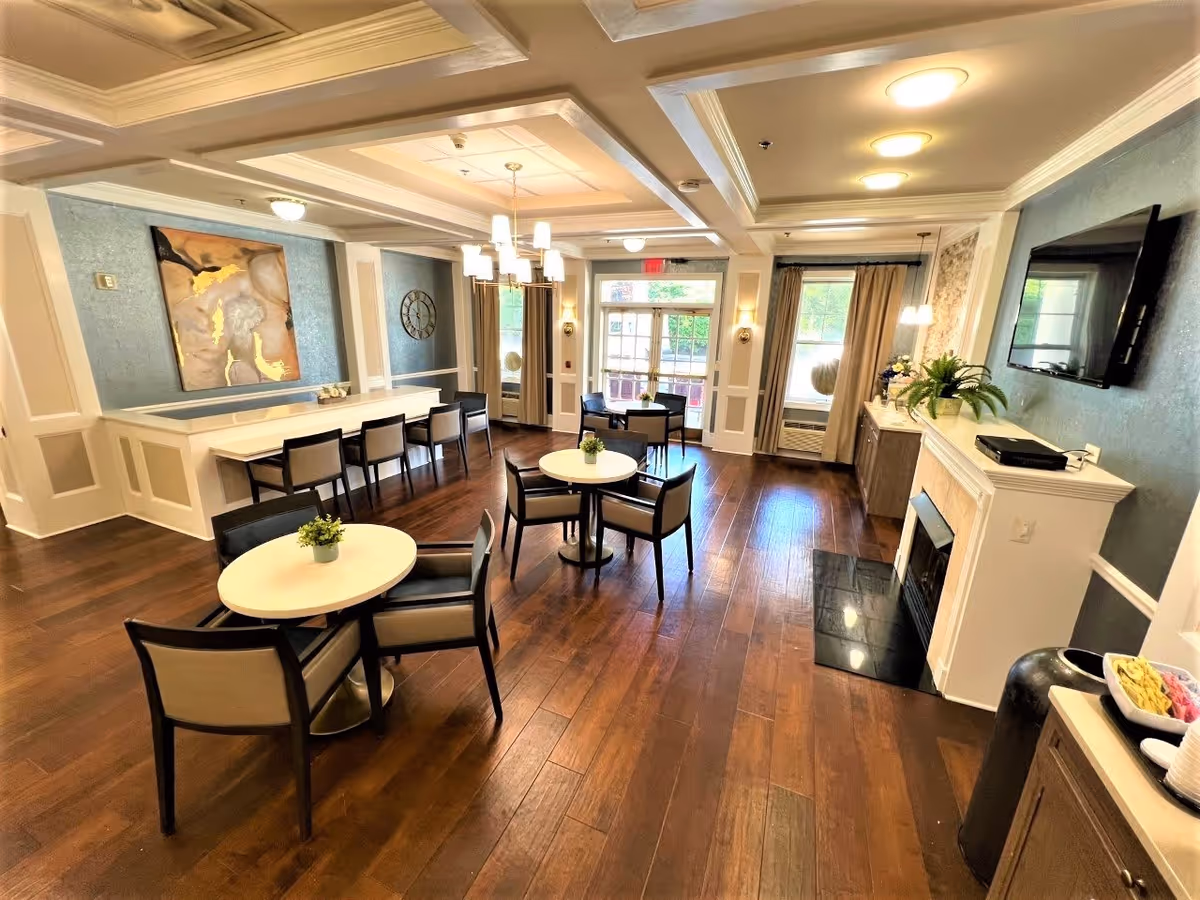 A bright and spacious common area in an assisted living facility featuring several round tables with chairs, a long counter with bar stools, a wall-mounted TV above a fireplace, decorative plants, artwork, and large windows with curtains allowing natural light to fill the room.