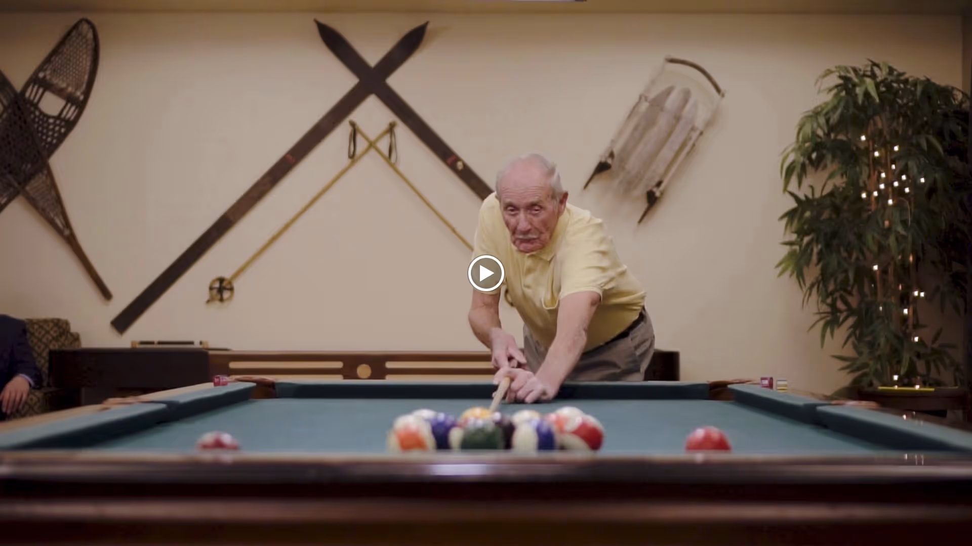 An elderly man in a yellow shirt is playing pool, aiming his cue stick at a rack of billiard balls on a pool table in a recreation room decorated with vintage skis, a sled, and a large plant with string lights.