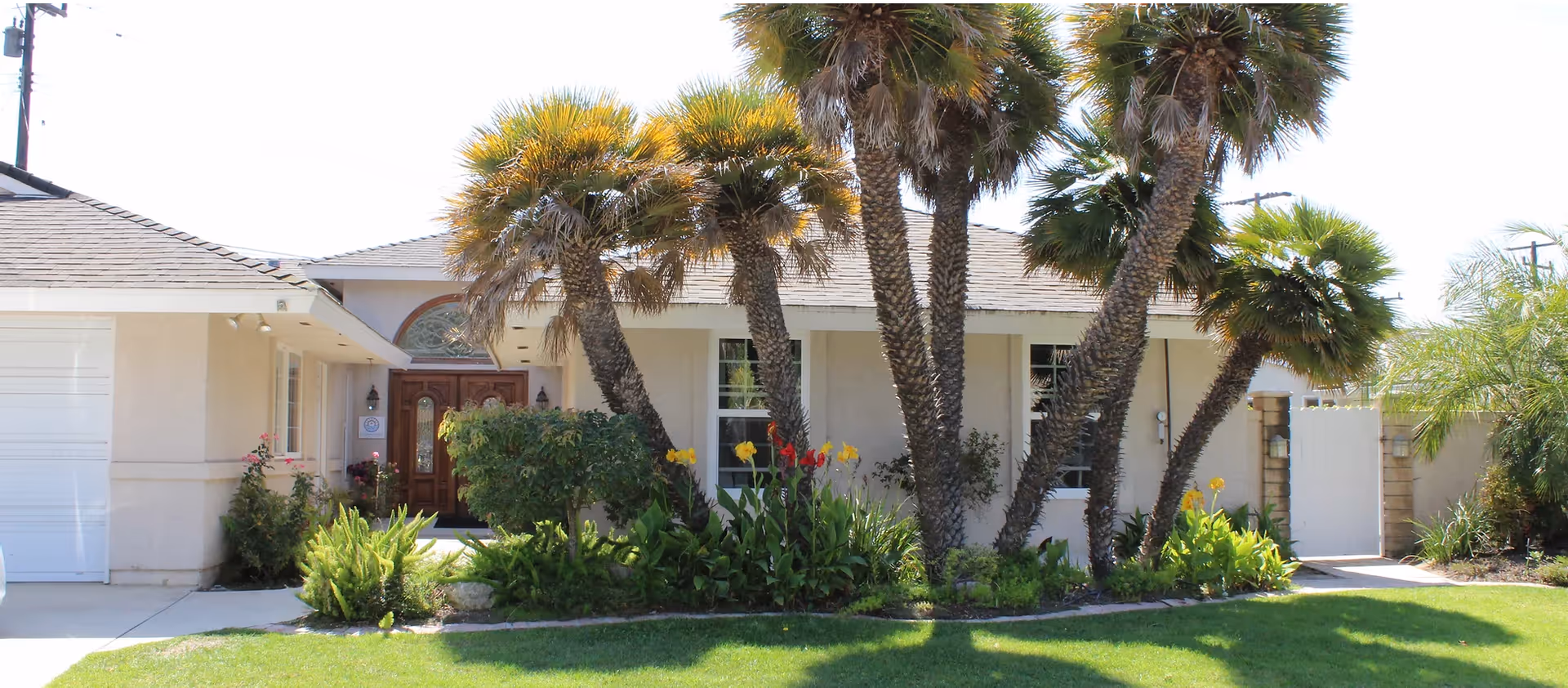 Front exterior view of a single-story building with a beige facade, a wooden double door entrance, and several palm trees and flowering plants in the landscaped garden in front. There is a white garage door on the left and a white gate on the right side of the building.