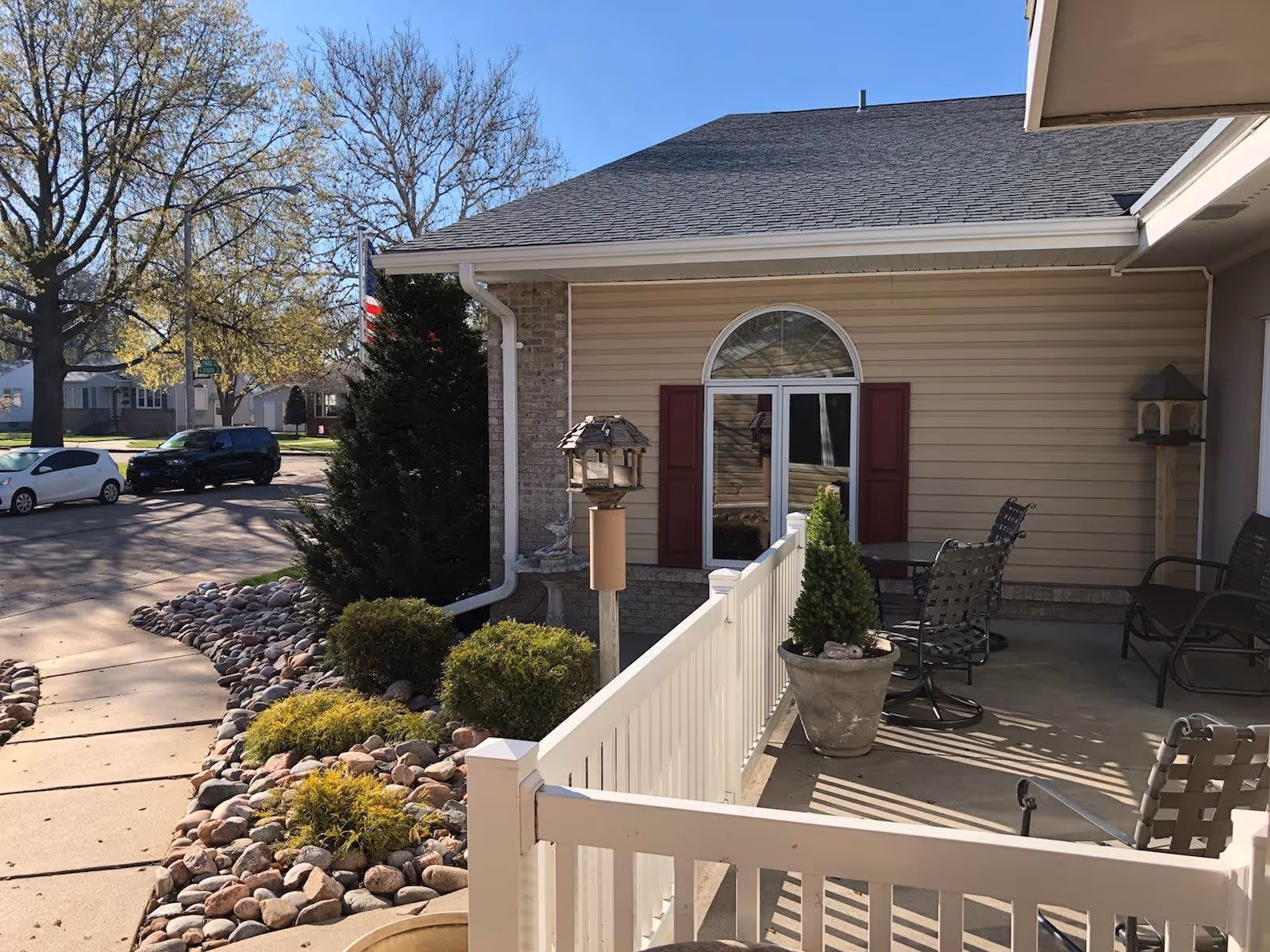 Outdoor patio area of a retirement home with a white railing, potted plants, metal chairs, and a small table. The building has beige siding with a window featuring red shutters and an arched window above. There are trees, parked cars, and a sidewalk visible in the background under a clear blue sky.