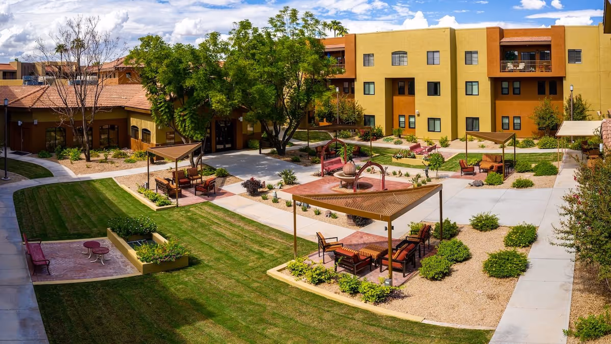 Outdoor courtyard area of a senior living facility with green lawns, paved walkways, shaded seating areas with chairs and tables, a central water fountain, and surrounding multi-story buildings under a partly cloudy blue sky.