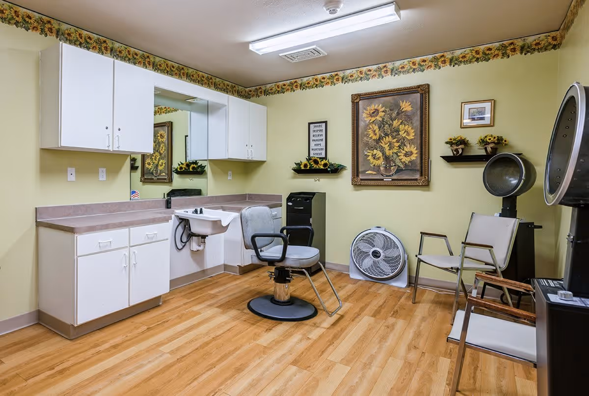 Interior of a hair salon room with a salon chair in front of a sink, white cabinets, a large mirror, two hair dryers, two chairs, a fan on the floor, and sunflower-themed wall decorations including a painting and small shelves with flower arrangements.