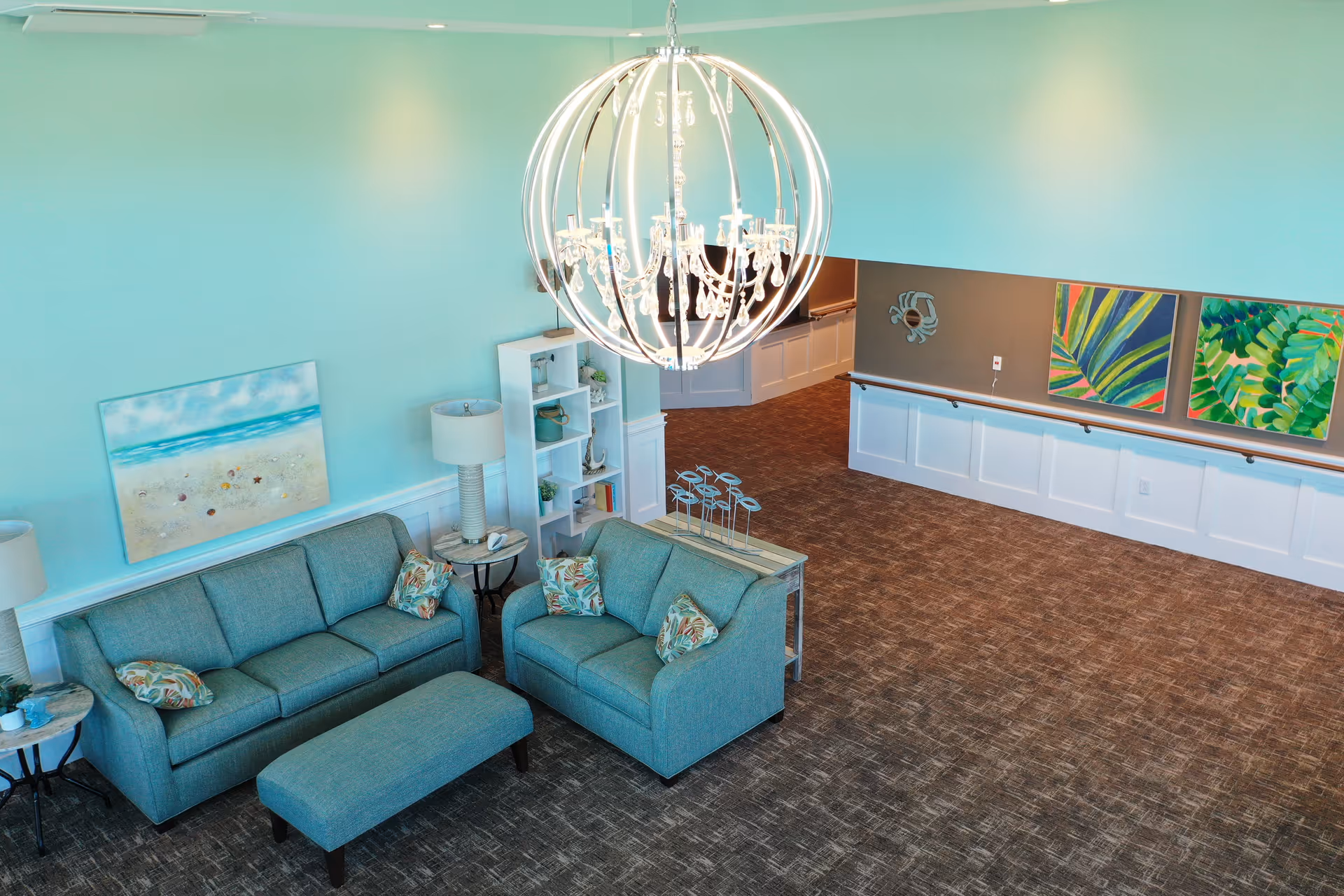 A bright living room with teal walls and carpeted floor. The room features a teal sofa, loveseat, and matching ottoman with floral patterned pillows. There are two side tables with lamps, a white bookshelf with decorative items, and a large, modern chandelier hanging from the ceiling. The far wall has two colorful leaf paintings and a decorative mirror.