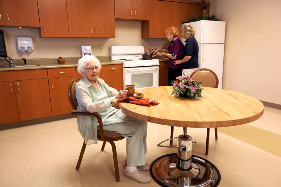 An elderly woman sitting at a round wooden table holding a cup in a kitchen area. Two other elderly women are standing near the stove, cooking together. The kitchen has wooden cabinets, a white stove, and a white refrigerator. A floral arrangement is on the table.