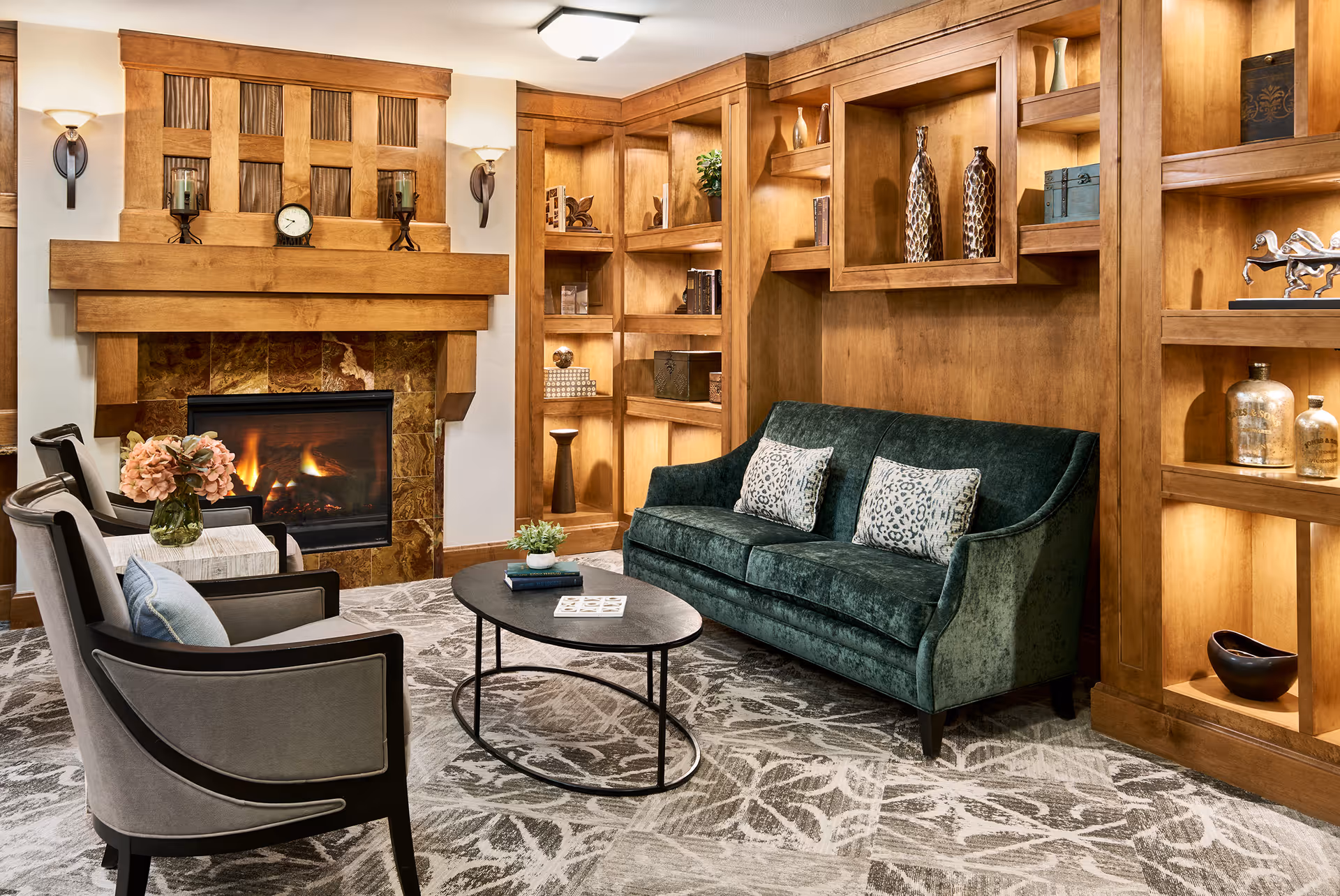 A cozy living room with a green velvet sofa adorned with two patterned pillows, two gray armchairs with light blue cushions, a black oval coffee table with books and a small plant, and a fireplace with a wooden mantel and decorative items. The room features built-in wooden shelves with various decorative objects and warm lighting.