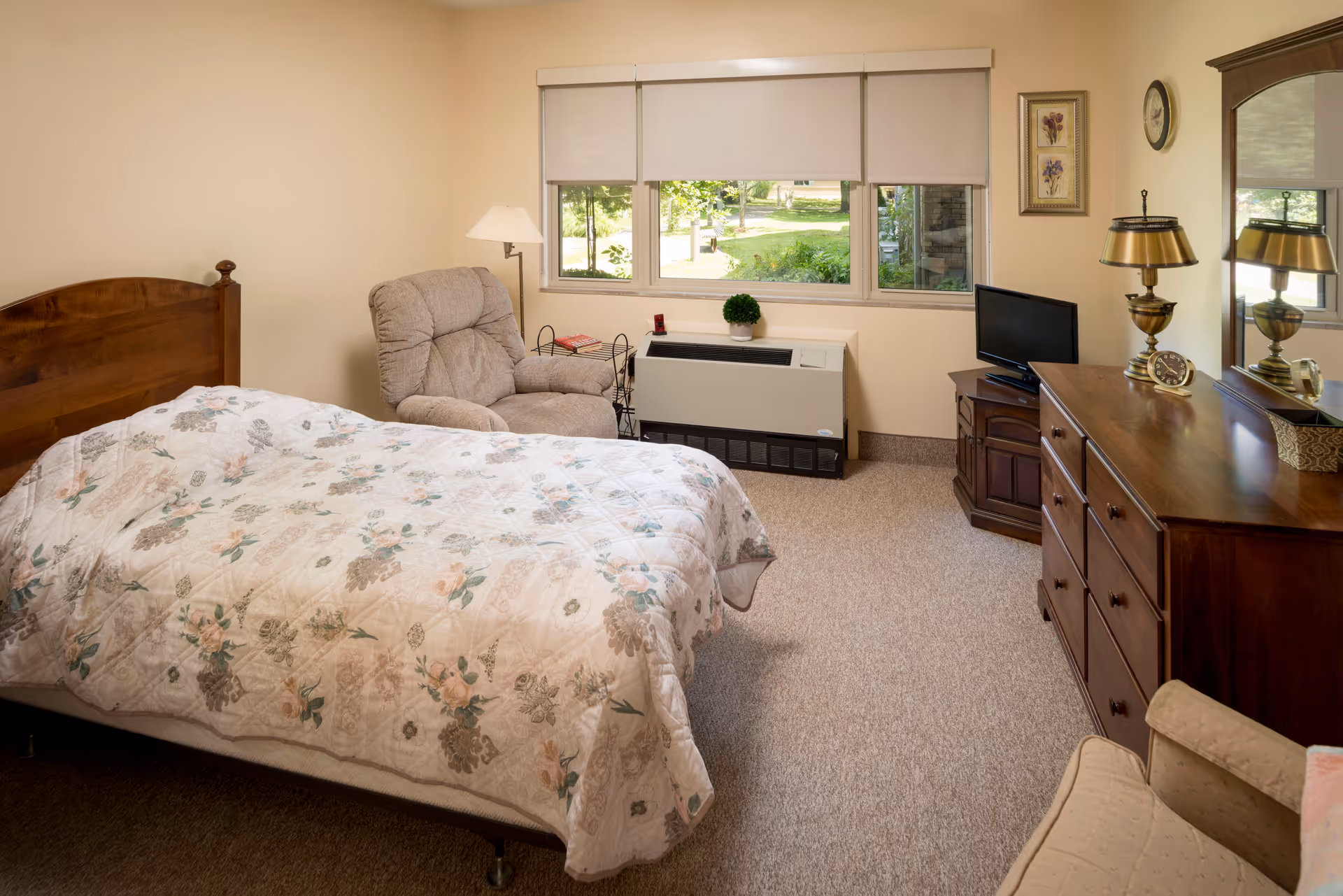 A cozy bedroom with a wooden bed frame and floral patterned bedspread, a beige recliner chair next to a window with white blinds, a wooden dresser with a mirror, a small TV on a corner cabinet, a brass lamp, a clock, and framed floral artwork on the wall. The window shows a green outdoor garden area.
