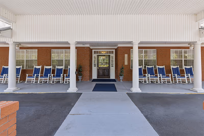 Covered front entrance of a brick senior living facility with white columns and rows of blue-cushioned rocking chairs.