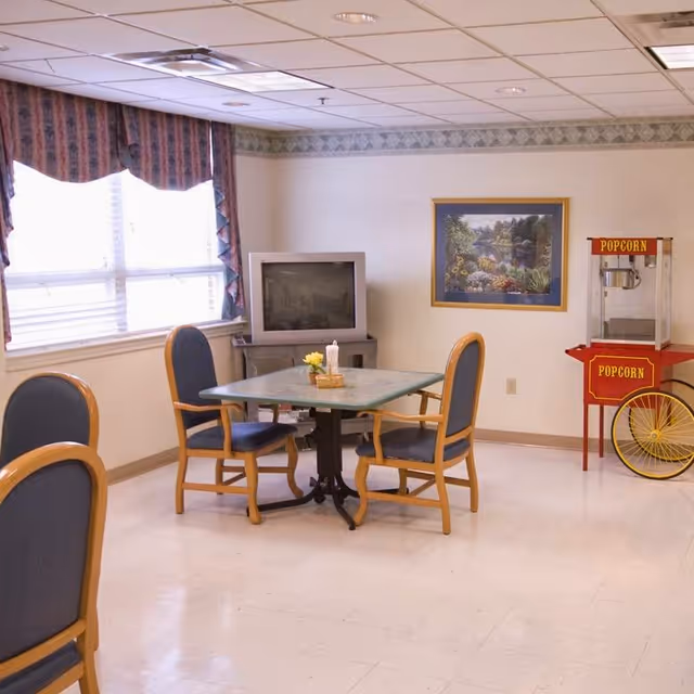 A small common area with a square table surrounded by four wooden chairs with blue cushions. A TV sits on a stand against the wall, next to a red popcorn machine on wheels. A framed landscape painting hangs on the wall above the popcorn machine. The room has a large window with patterned curtains and a tiled floor.