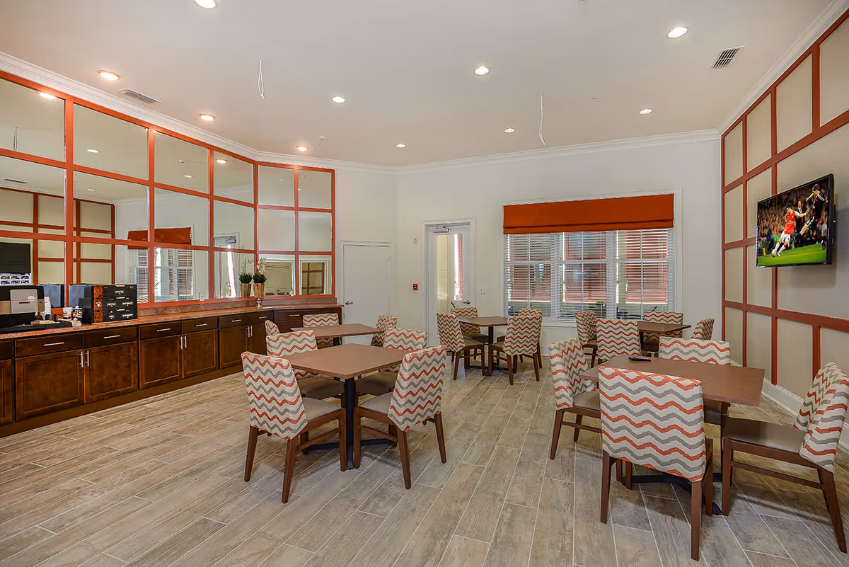 Communal dining room with multiple tables and zigzag-patterned upholstered chairs, a wall-mounted TV, and mirrored cabinetry along one wall.