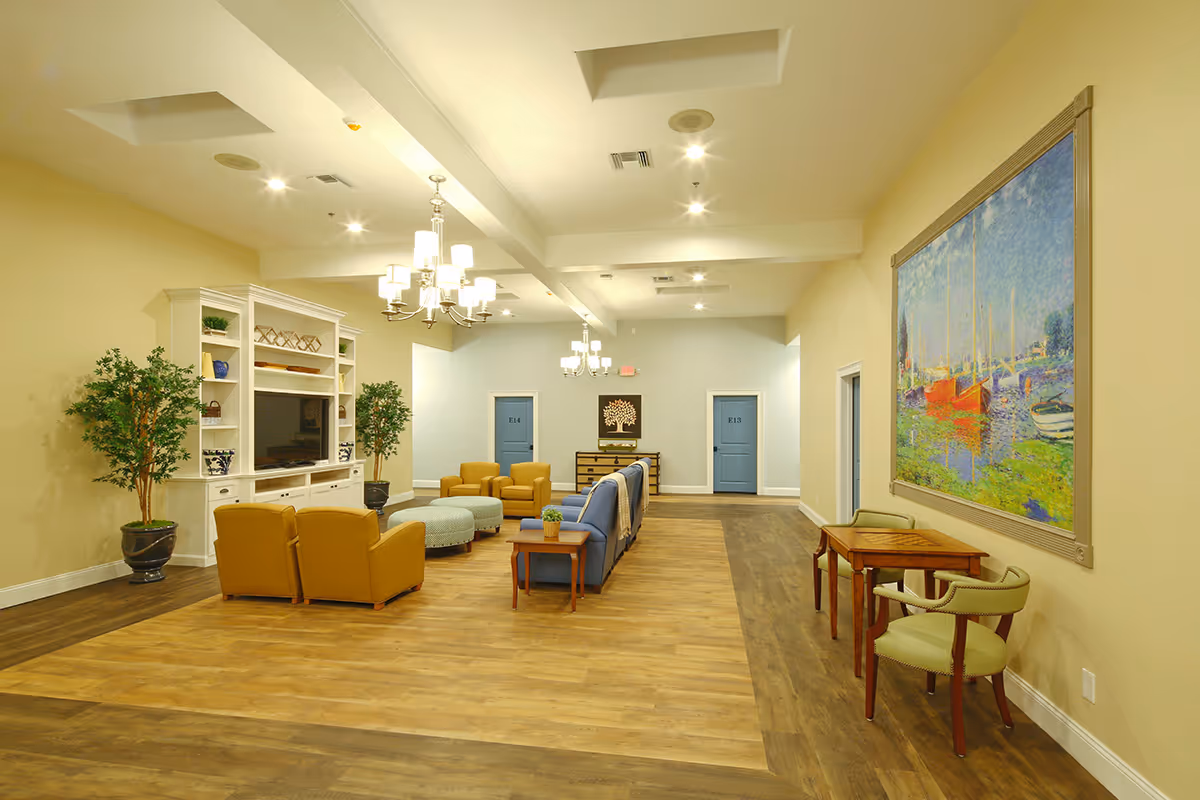 A spacious and well-lit living room area in an assisted living facility featuring comfortable seating including yellow armchairs and a blue sofa, a large white entertainment center with a TV, two potted plants, a wooden side table with a small plant, a small table with two green chairs, and a large colorful painting of boats on the wall. The room has wooden flooring and blue doors labeled E13 and E14 in the background.