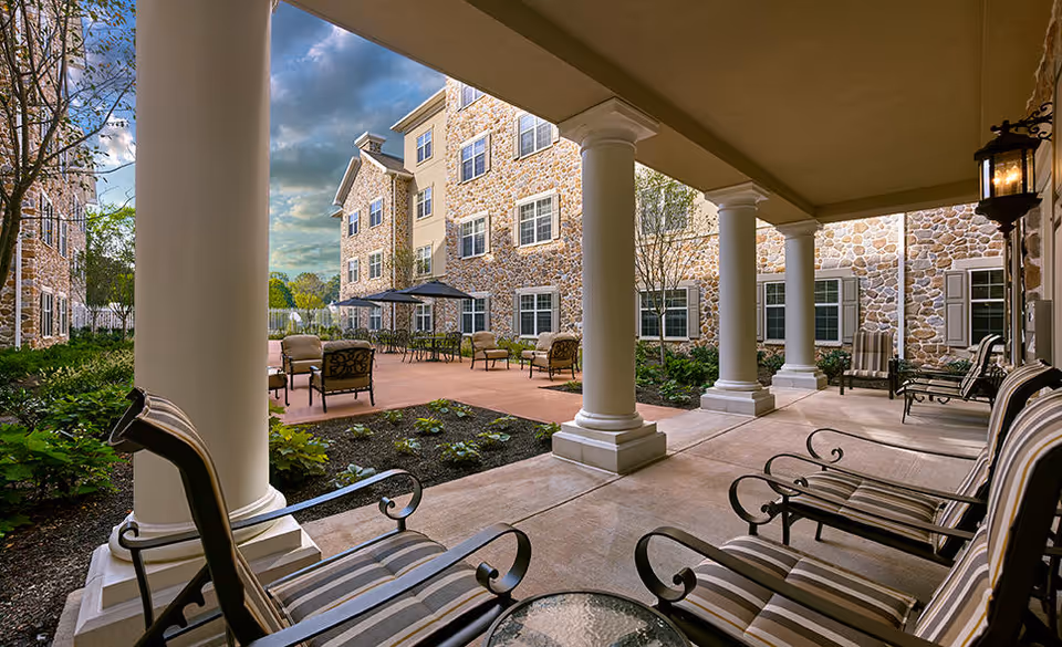 Covered porch with striped cushioned chairs and columns overlooking a landscaped courtyard and stone-faced senior living building.