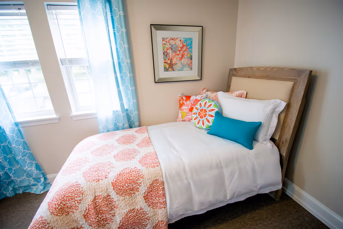 A neatly made single bed with a wooden headboard in a bedroom. The bed has white sheets, a coral-patterned quilt, and several colorful decorative pillows including turquoise and coral colors. There is a framed artwork on the wall above the bed and two windows with blue patterned curtains letting in natural light.