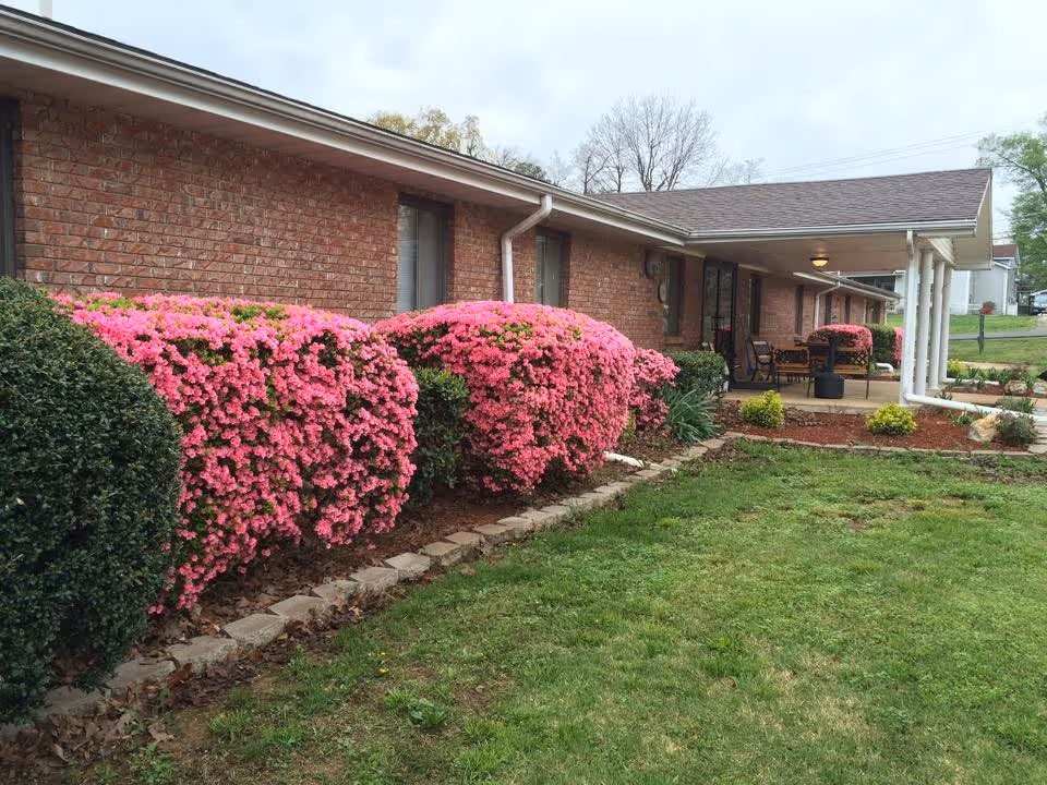 Exterior view of a single-story brick building with a covered porch area. The porch has outdoor seating and is surrounded by well-maintained landscaping, including vibrant pink flowering bushes and green shrubs. The lawn in front is green and neatly trimmed, and the sky is overcast.