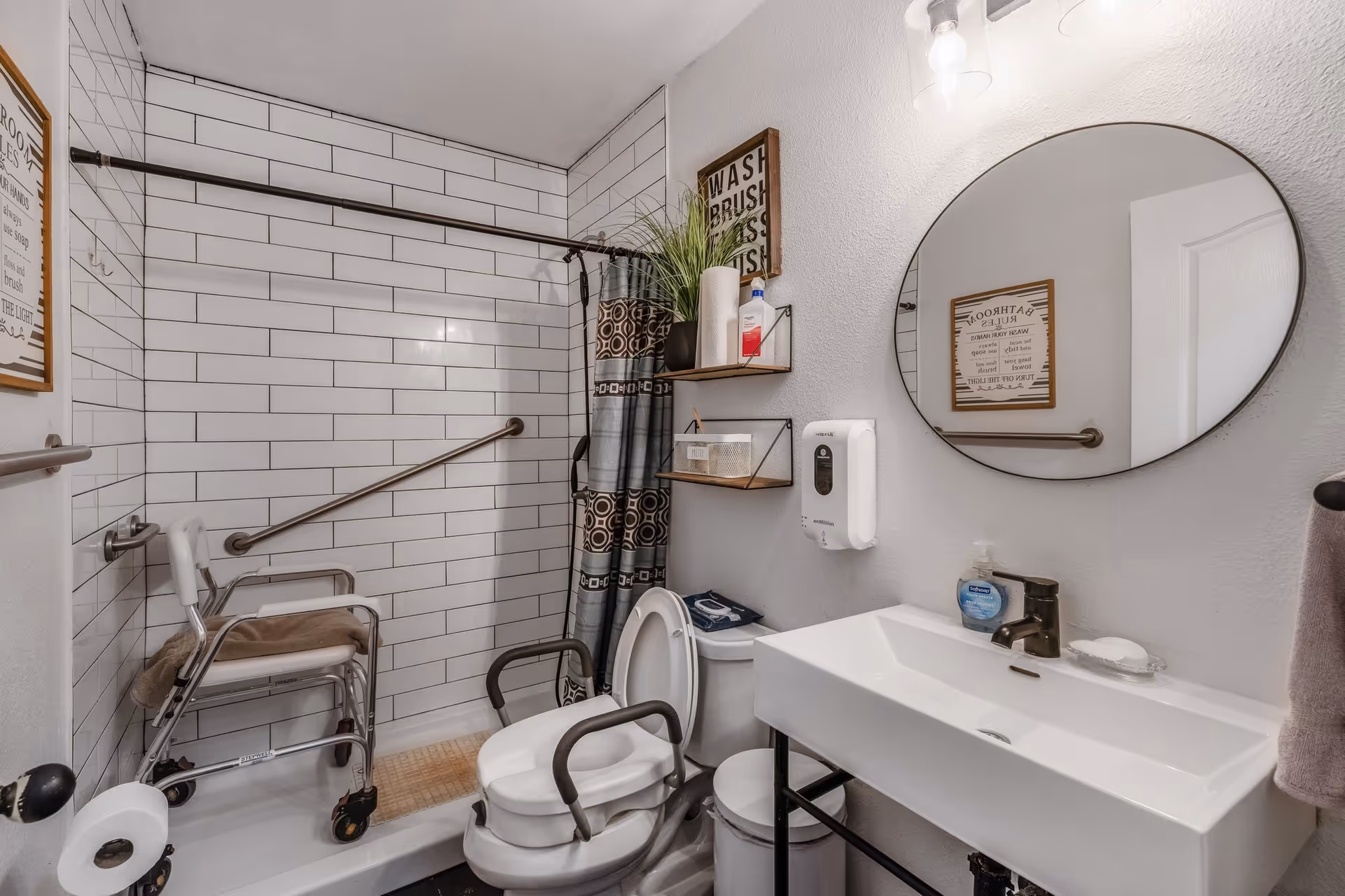 Accessible bathroom with a tiled walk-in shower, shower chair and grab bars, a raised toilet seat, sink and round mirror.