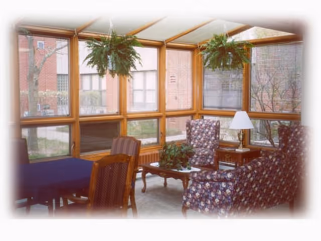 A sunroom with large windows showing an outdoor view of trees and a brick building. Inside, there are two floral upholstered armchairs, a wooden coffee table with a plant, a side table with a lamp, and a dining table with wooden chairs. Two hanging plants are suspended from the ceiling.
