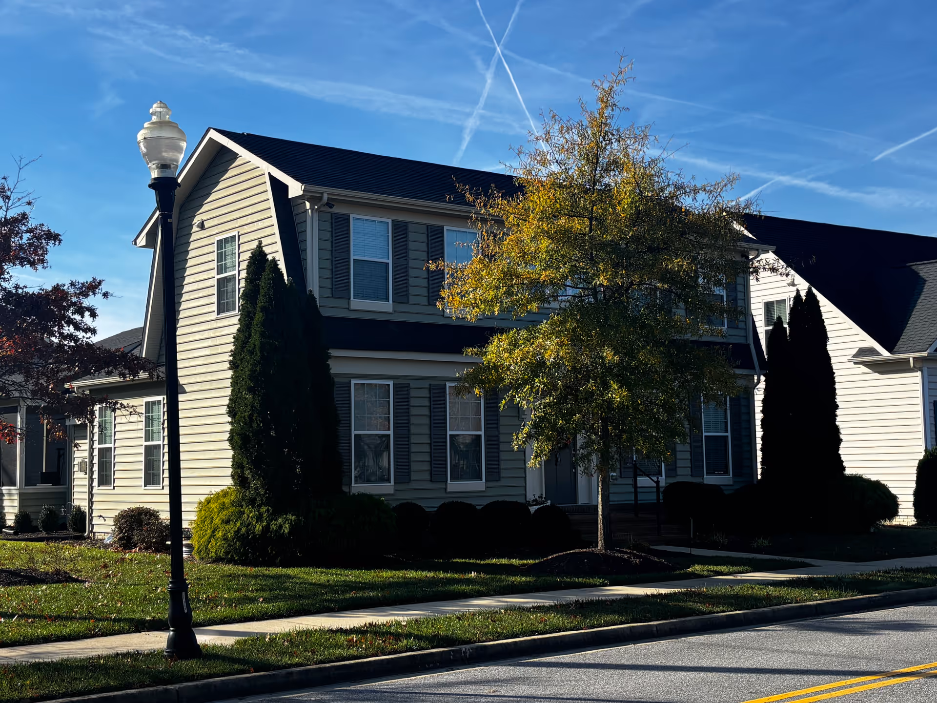 Exterior view of a two-story residential building with beige siding and multiple windows. There is a sidewalk and a street lamp in front, along with a tree and some bushes in the landscaped yard. The sky is clear with visible contrails.