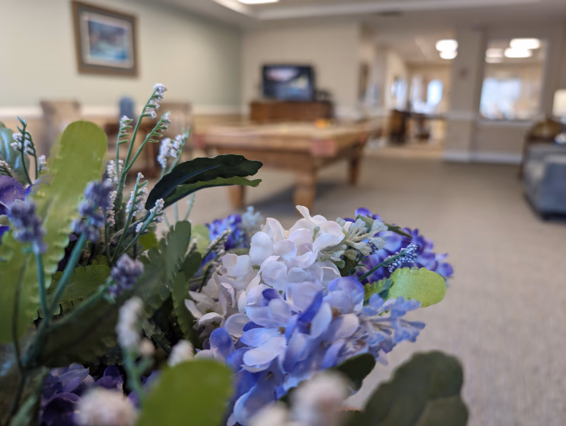 Close-up of a bouquet of artificial flowers with purple and white petals in the foreground, with a blurred background showing a room with a pool table, chairs, and a television.