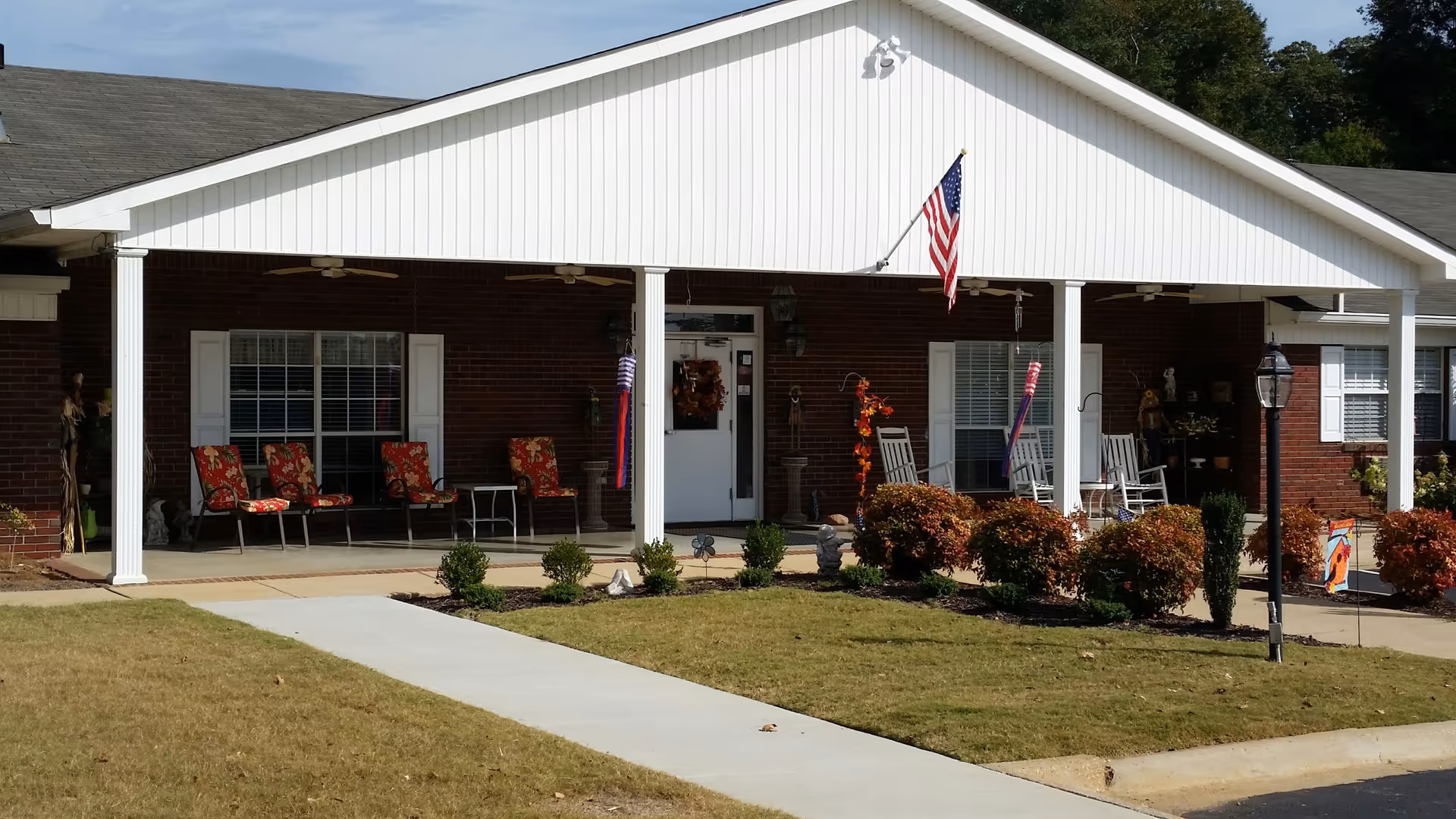 Front exterior view of a single-story brick building with a white porch roof supported by white columns. The porch has several chairs with red floral cushions and white rocking chairs. An American flag is mounted near the entrance door, which has a fall-themed wreath. There are small bushes and a lawn in front of the building.