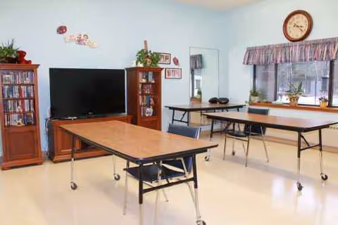 An activity room with rolling tables, chairs, a TV, bookshelves, and a wall clock.