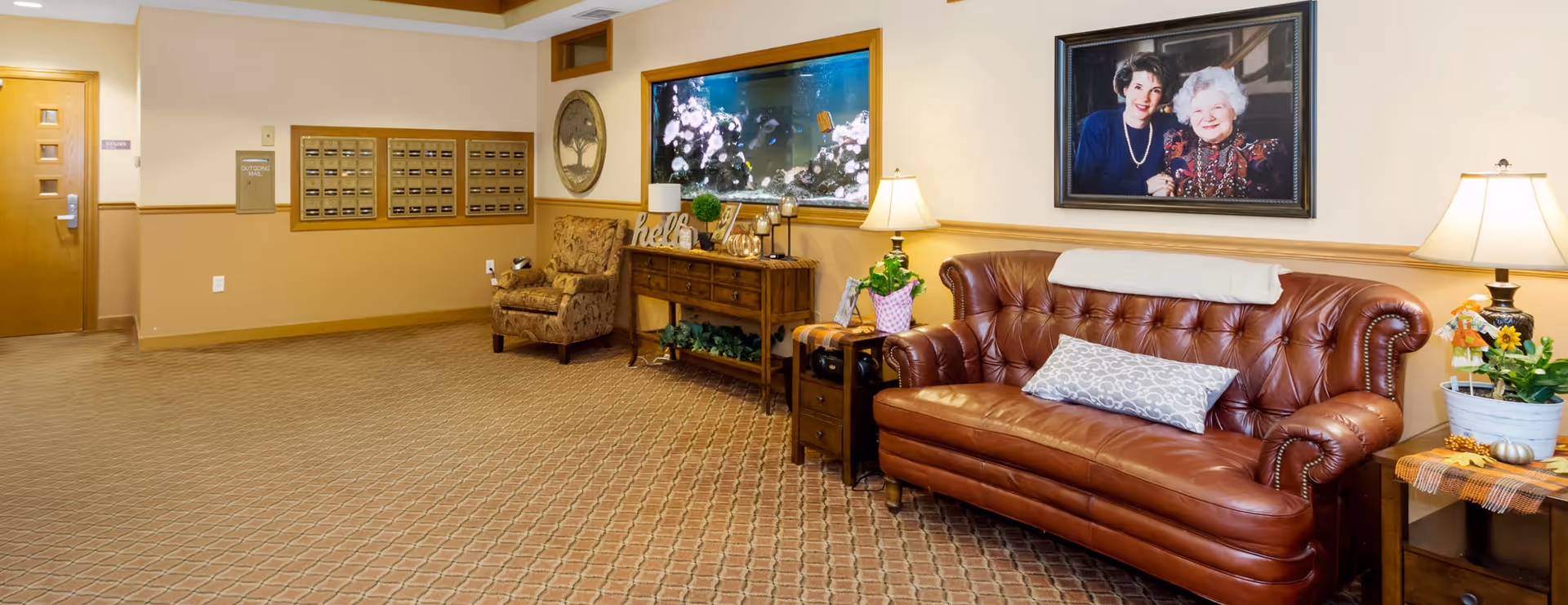 A cozy seating area in a senior living facility with a brown leather couch, a patterned armchair, two side tables with lamps and plants, a wooden console table with decorative items, a large aquarium built into the wall, and a framed photograph of two women hanging above the couch. The floor is carpeted and there are mailboxes mounted on the wall near a wooden door.