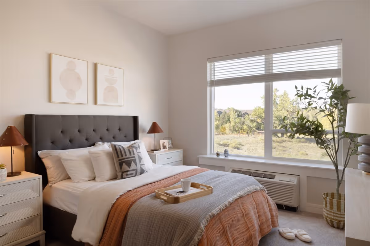 A cozy bedroom with a large window showing an outdoor view with trees. The room features a bed with a tufted dark headboard, white and patterned pillows, and a layered blanket in shades of gray and orange. There are two bedside tables with matching lamps, a small plant, a framed photo, and a tray with a cup on the bed. A potted plant stands near the window, and slippers are placed on the carpeted floor.