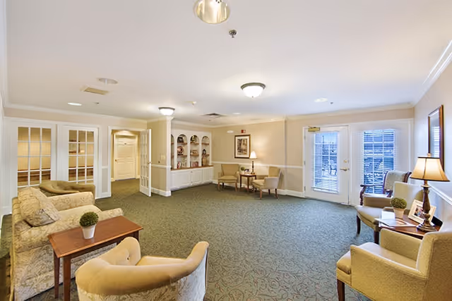 A spacious and well-lit living room area with beige walls and carpeted floor. The room features several upholstered armchairs and sofas arranged around small wooden tables with decorative plants and lamps. French doors with blinds lead outside, and built-in shelves with decorative items are visible along one wall.