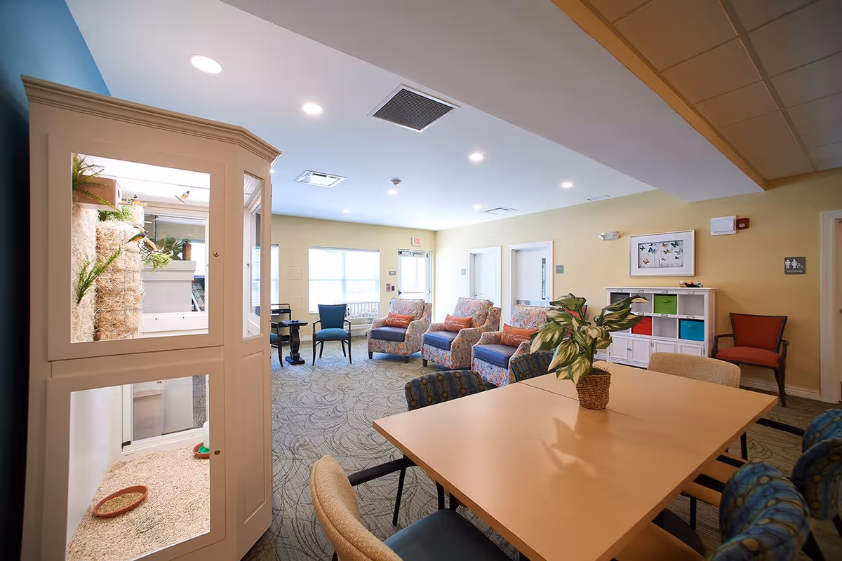 A bright and spacious common area in a senior living facility with a table and chairs in the foreground, a potted plant on the table, several patterned armchairs with orange pillows, a white storage unit with colorful bins, and a small indoor enclosure with plants and birds on the left side.