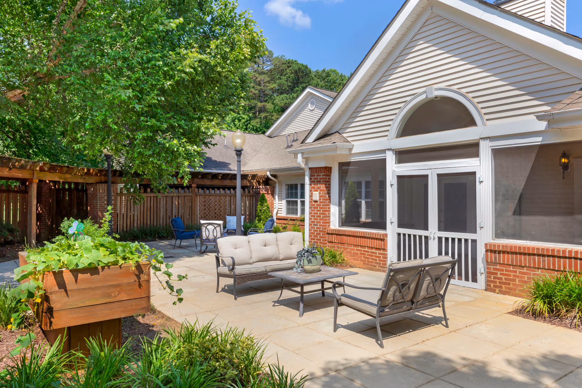 Outdoor patio area at Brookdale Cary featuring cushioned metal seating with a sofa and two chairs around a coffee table, a raised wooden planter with greenery, a large tree providing shade, and a screened porch attached to a brick and siding building under a clear blue sky.