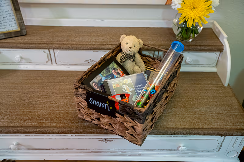 A woven basket labeled 'Sharon' sits on a wooden dresser. Inside the basket are a small teddy bear with a bow tie, a framed photo, a music CD, a tube with colorful beads, and a small red item. A vase with yellow and white flowers is placed on the dresser next to the basket.