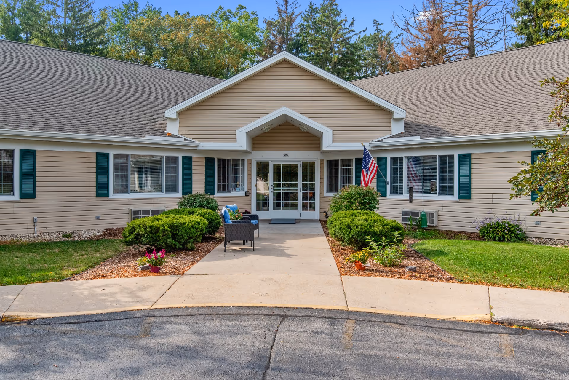 Front entrance of a single-story senior living building with a covered entry, seating, American flag, and landscaped walkway.