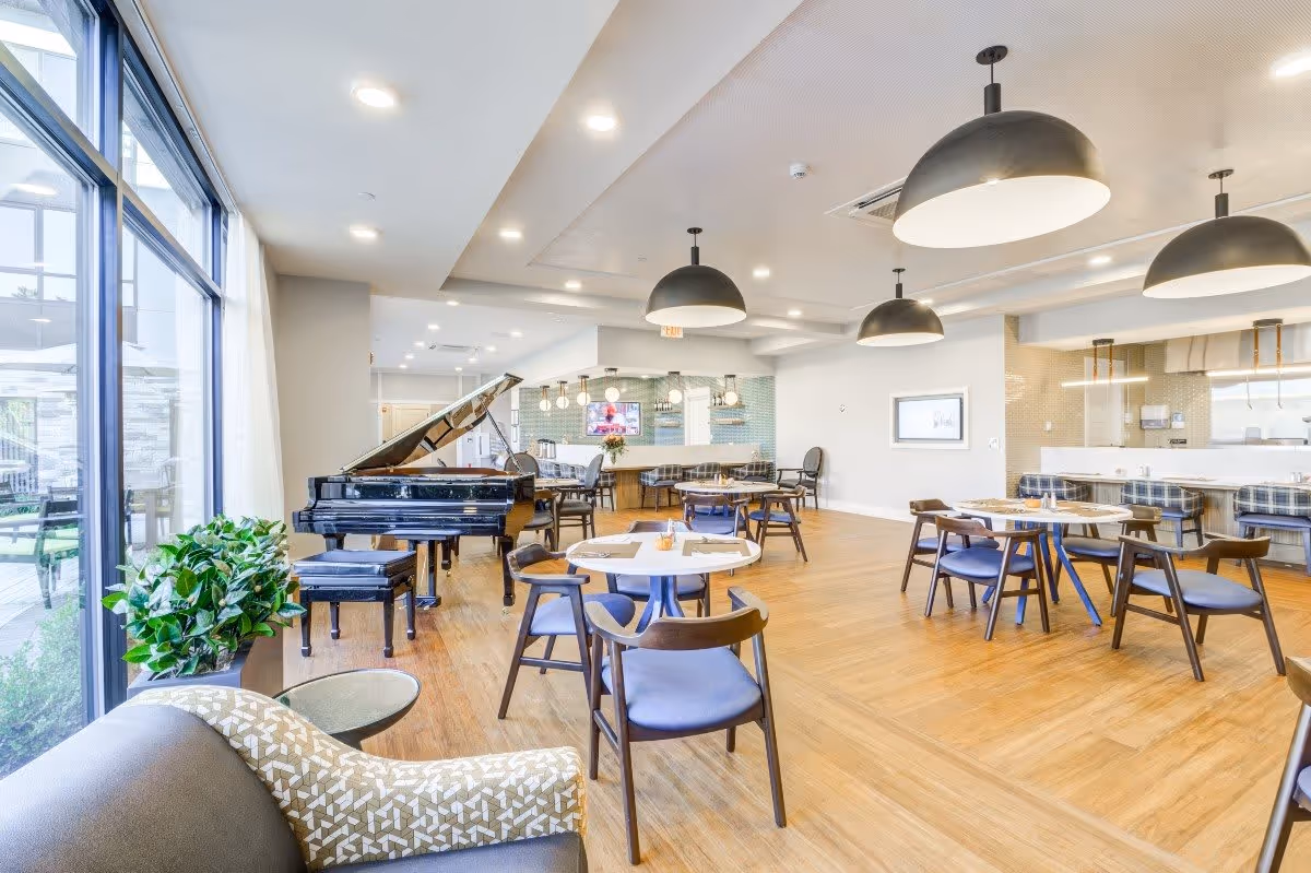 Bright communal dining and lounge area with round tables and chairs, a grand piano, large windows, and pendant lights.