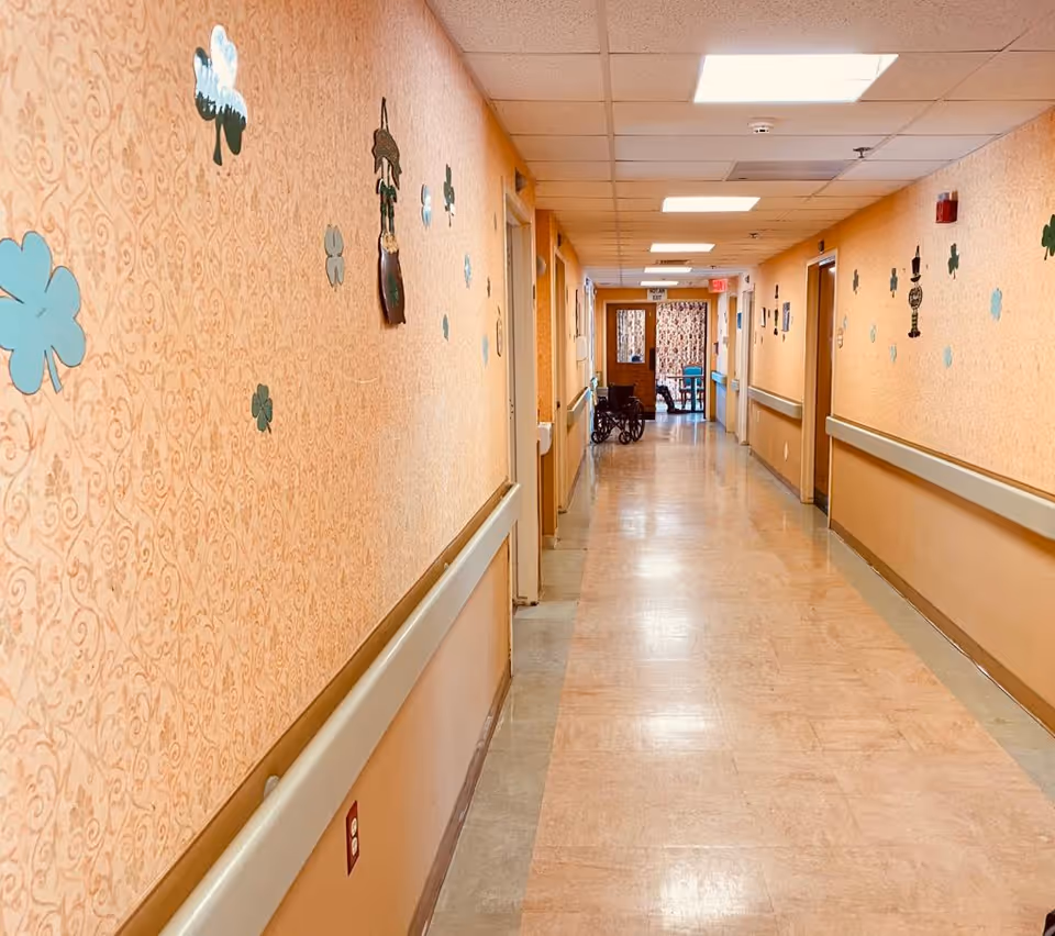 A long, well-lit hallway in a senior living facility with beige patterned walls decorated with green clover and other small wall hangings. There are handrails along both sides of the hallway, several closed doors, and a wheelchair positioned near the far end. The floor is a light-colored linoleum, and the ceiling has recessed lighting panels.