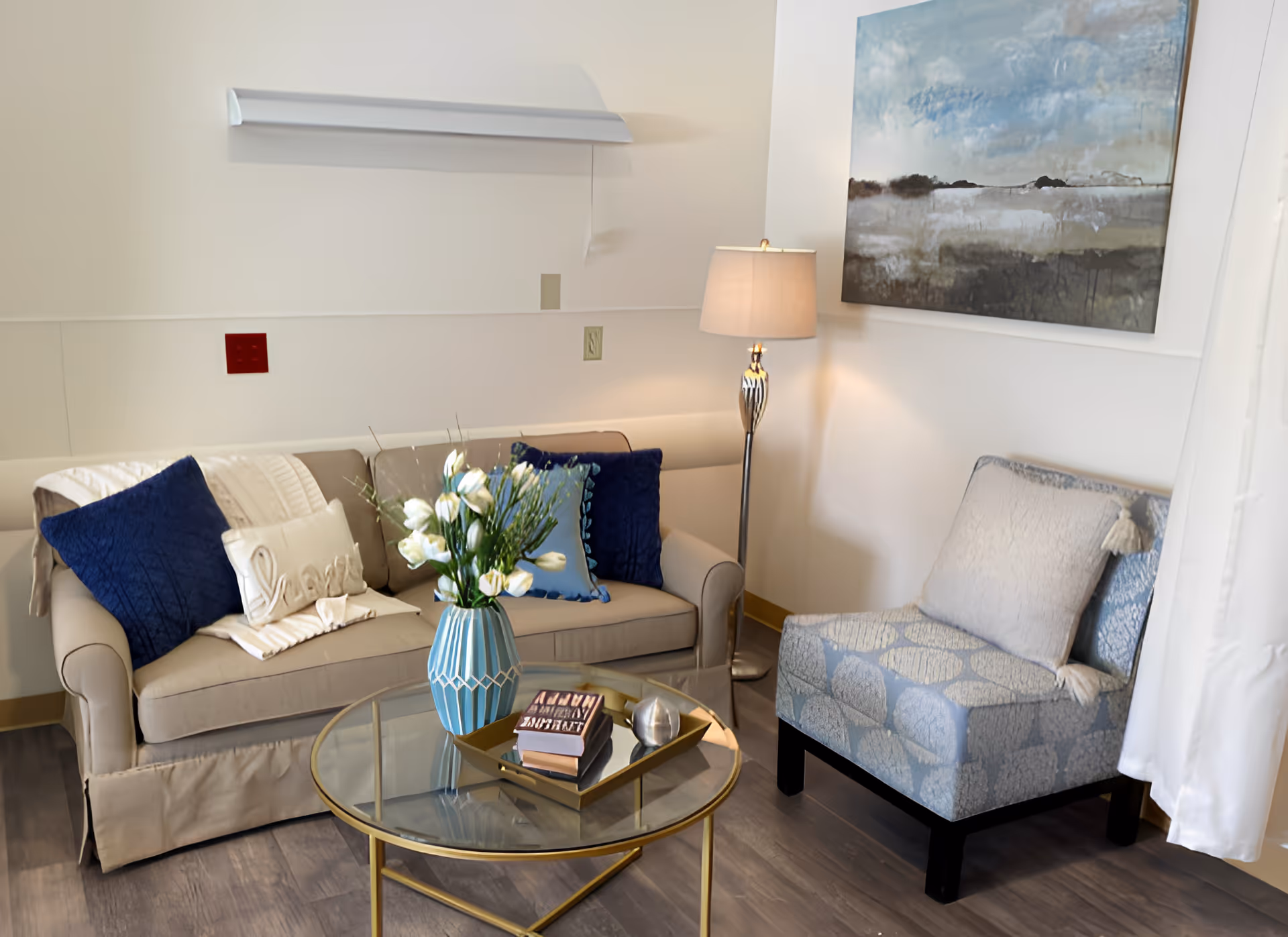 A cozy living room area with a beige sofa adorned with blue and white pillows, a glass coffee table with a blue vase of white flowers and books, a patterned blue armchair, a floor lamp, and a large abstract painting on the wall.