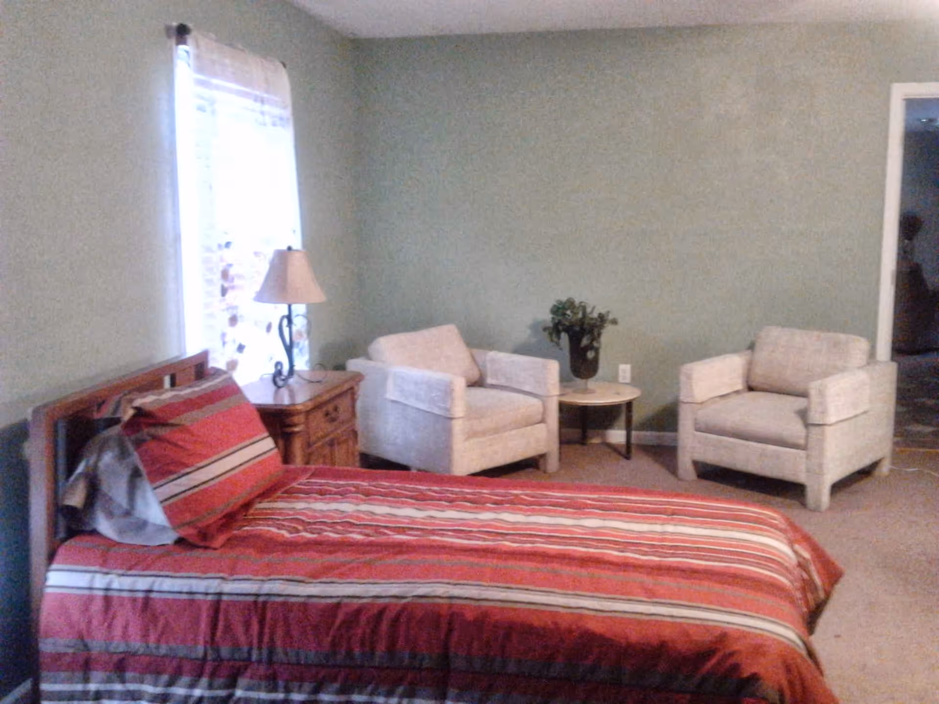 A bedroom with a single bed covered in a red and striped bedspread, a wooden nightstand with a lamp, two beige armchairs, and a small round table with a plant on it. There is a window with sheer curtains letting in natural light.