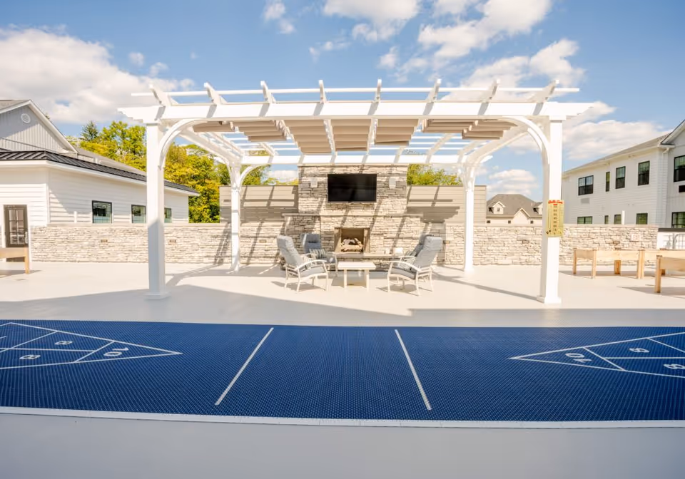 Outdoor patio area with a white pergola covering a seating arrangement of four cushioned chairs around a glass-top table in front of a stone fireplace with a mounted TV. The foreground features a blue shuffleboard court, and there are buildings and trees in the background under a partly cloudy sky.