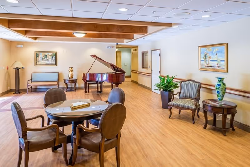 A spacious senior living common area with wooden flooring, featuring a round table with four cushioned chairs, a grand piano, a bench, two framed paintings on the walls, a potted plant, and a small side table with a decorative vase.