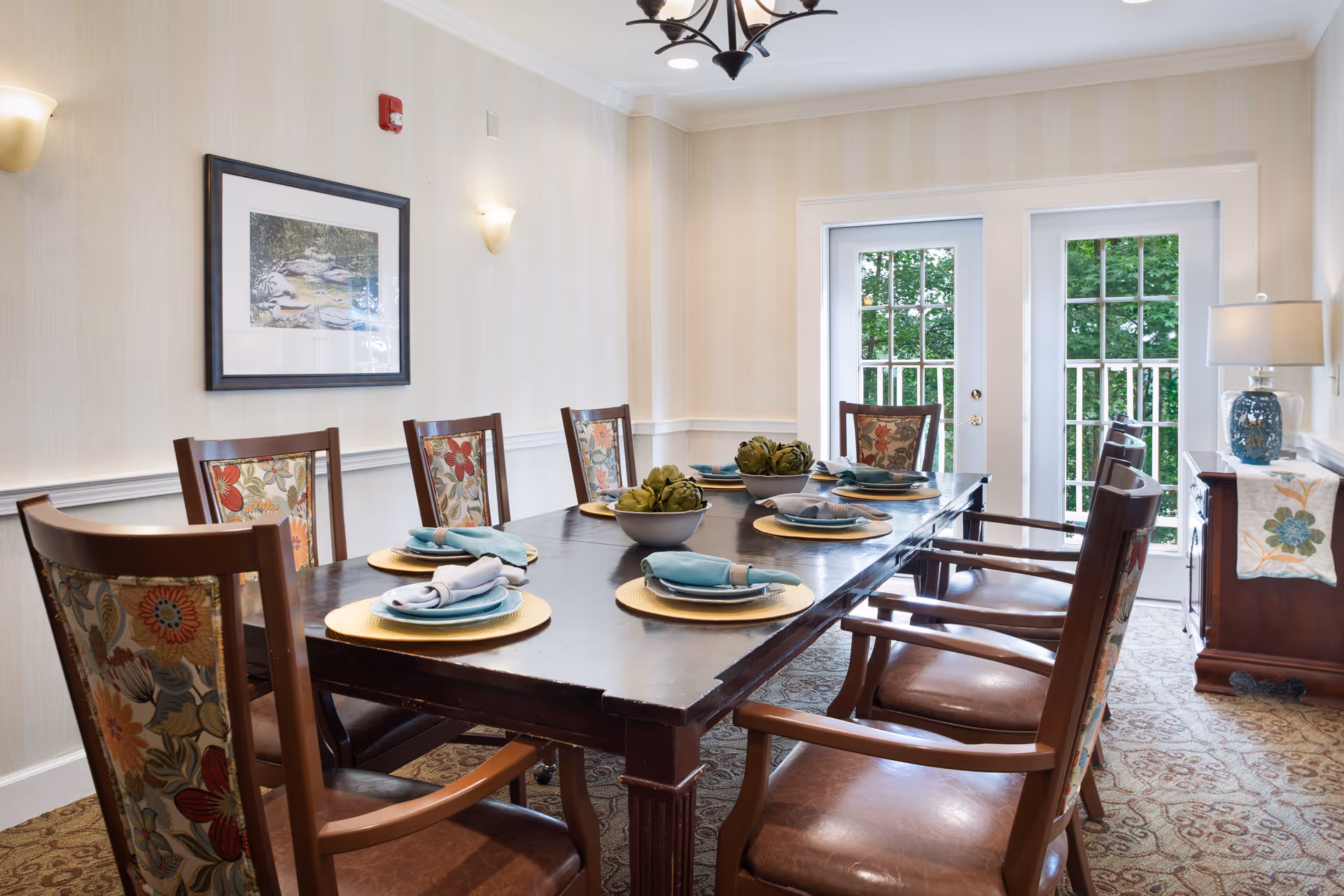 Dining room with a long dark wood table set with plates and napkins, floral-upholstered chairs, and French doors opening to greenery.
