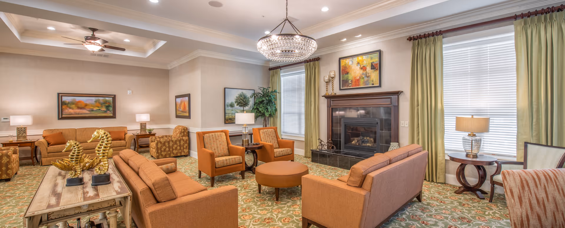 A spacious and well-lit living room with multiple orange upholstered sofas and armchairs arranged around a round ottoman. The room features a fireplace with a wooden mantel, decorative paintings on the walls, green patterned carpet, and large windows with green curtains. There are several side tables with lamps and decorative items including golden seahorse sculptures on a wooden table.