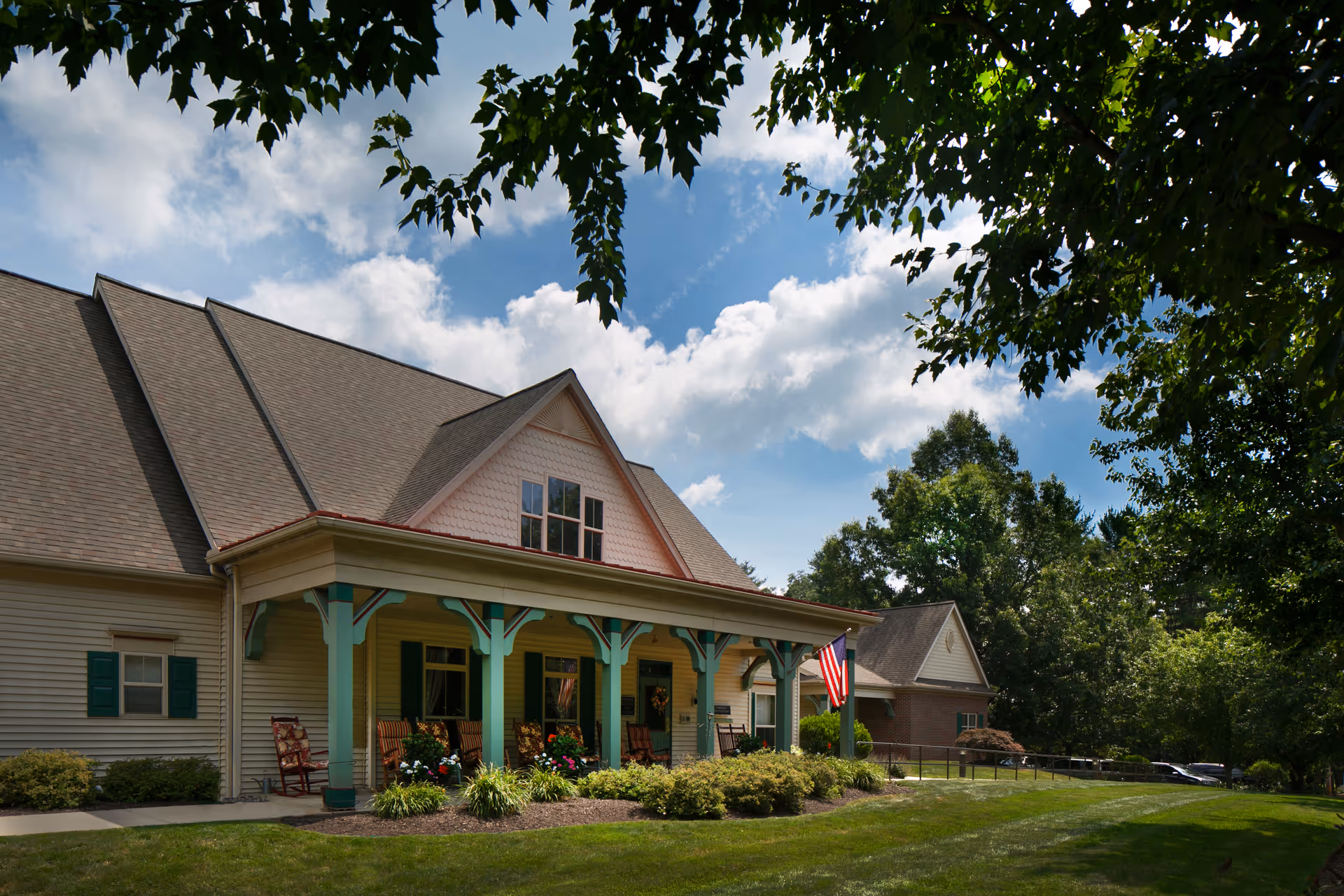 Exterior view of Columbia Cottage-Collegeville, a residential building with a large porch featuring green pillars, rocking chairs, and an American flag. The building is surrounded by green grass, bushes, and trees under a partly cloudy blue sky.