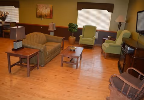 A cozy living room with wooden flooring, featuring a brown patterned sofa, two green armchairs, a wooden coffee table with a decorative bowl, side tables with lamps, a rocking chair, a flat-screen TV mounted on the wall, and a window with blinds and a valance.