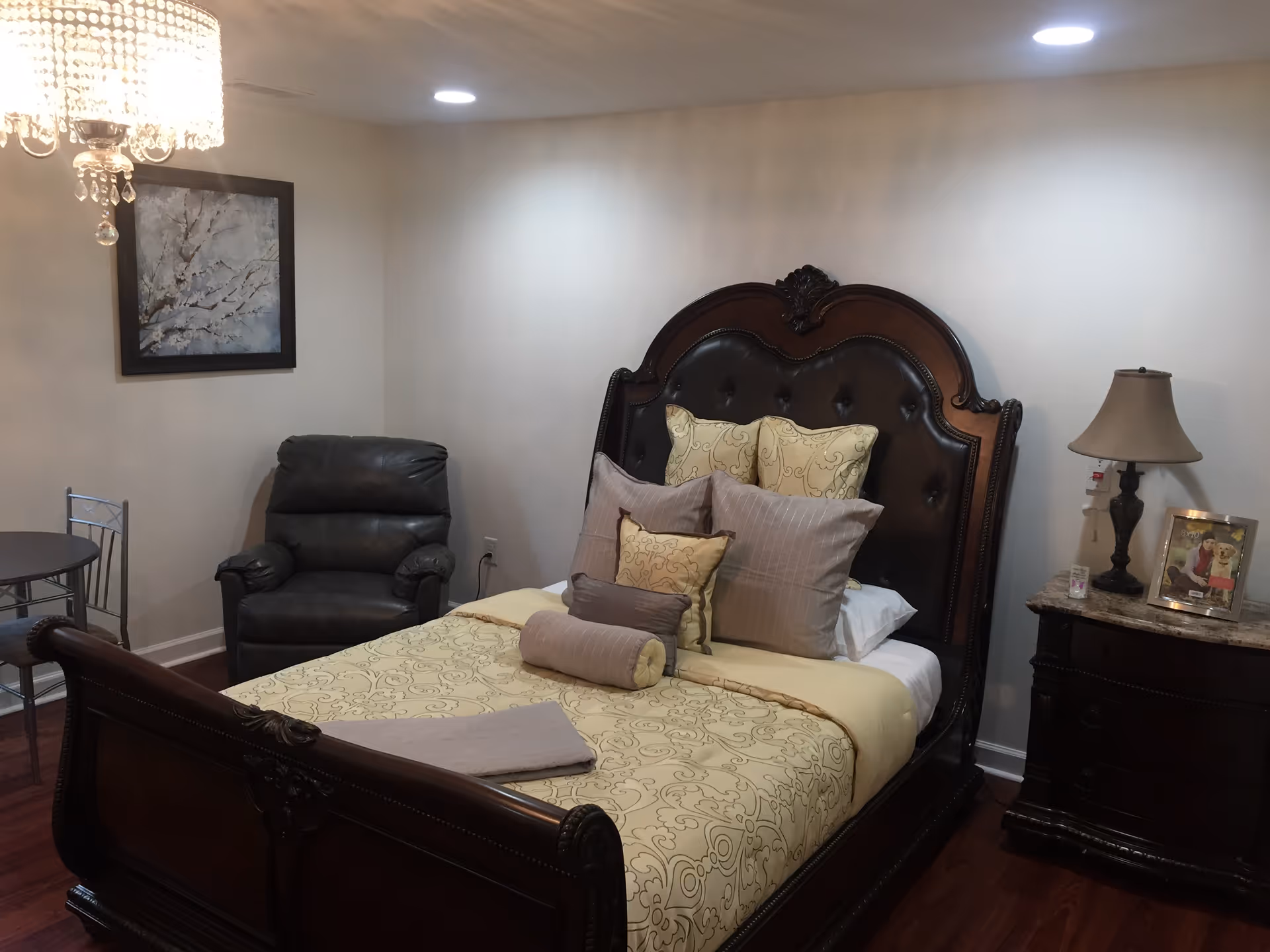A cozy bedroom with a dark wooden bed frame featuring an ornate headboard and footboard. The bed is neatly made with a yellow and beige patterned comforter and multiple pillows. To the right of the bed is a dark wooden nightstand with a lamp, a framed photo, and a candle. On the left side of the room, there is a black leather recliner chair and a small round table with two metal chairs. A chandelier hangs from the ceiling, and a framed artwork is mounted on the wall above the recliner. The room has wooden flooring and white walls.