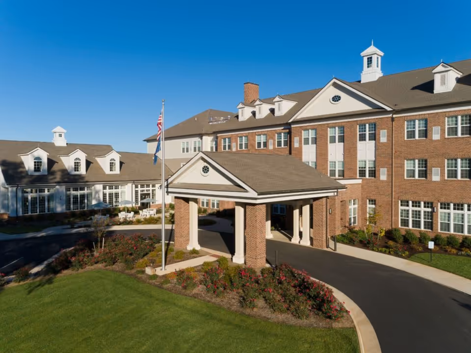 Exterior view of a large brick senior living facility building with multiple windows, a covered entrance supported by brick columns, a flagpole with the American flag, and well-maintained landscaping including green grass and flower beds under a clear blue sky.