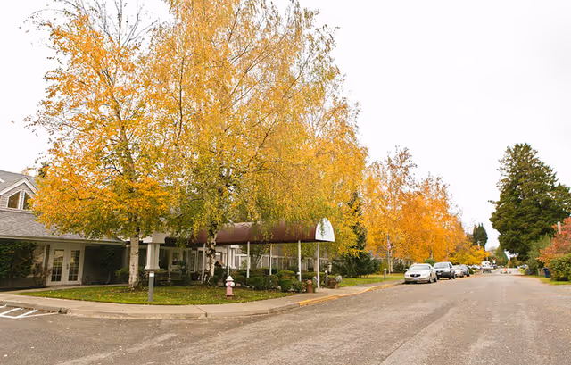 Street view of a senior living facility with a covered entrance, surrounded by trees with yellow autumn leaves. Several cars are parked along the road, and the building is partially visible behind the trees.