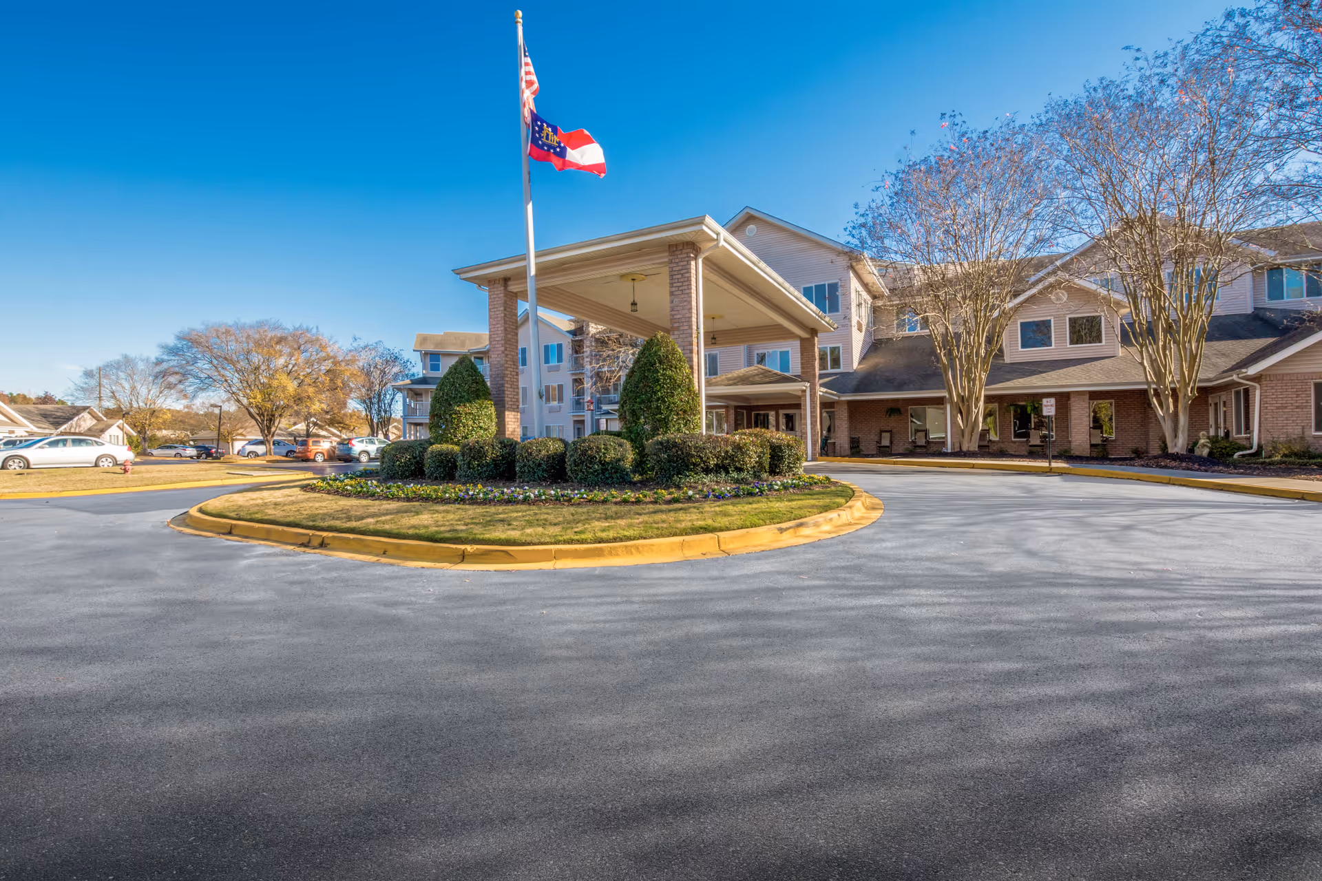 Exterior view of Washington Commons by Barclay House, showing a circular driveway with landscaped greenery and two flagpoles flying the American and Georgia state flags in front of a multi-story senior living facility building under a clear blue sky.