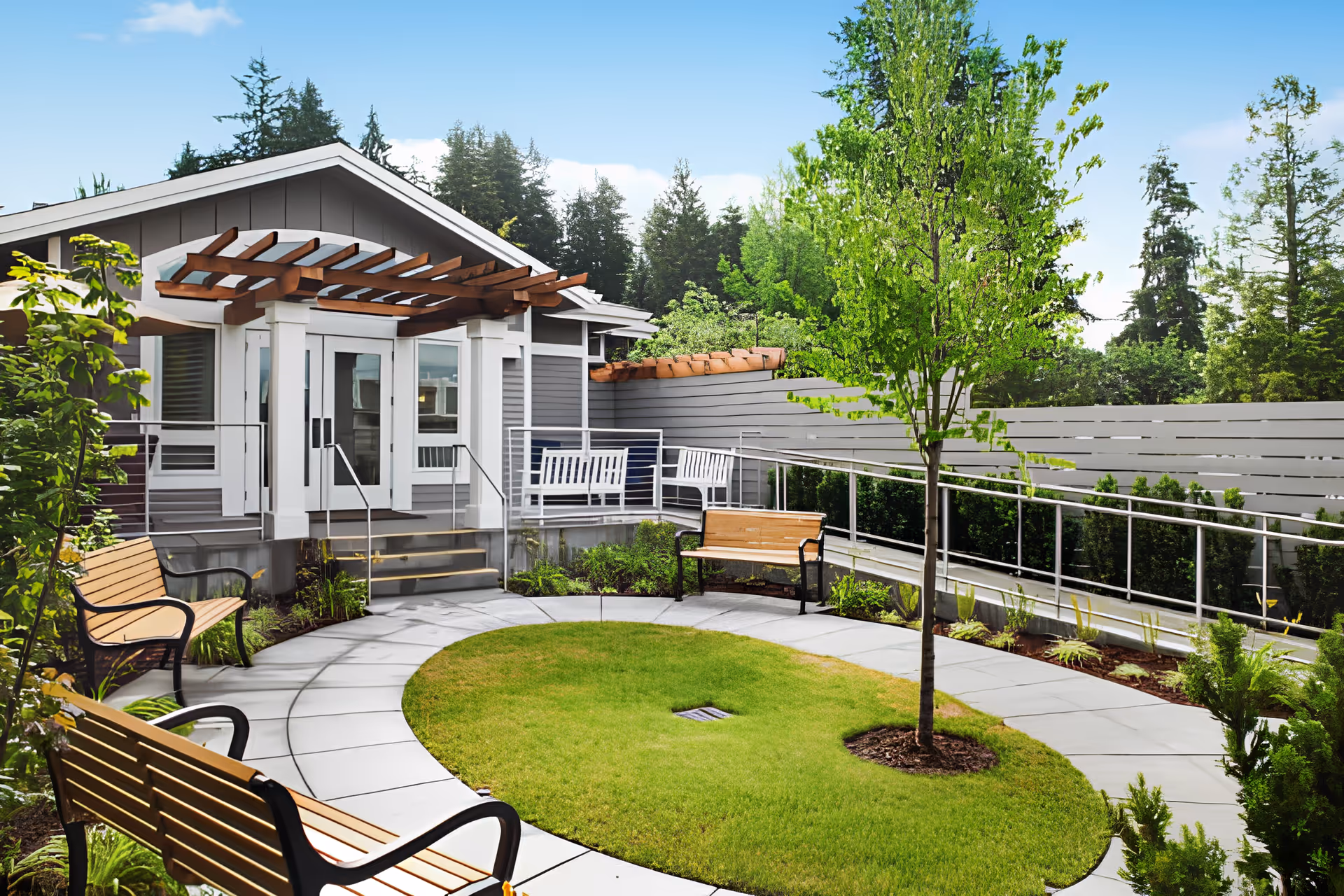 Landscaped courtyard with a circular lawn, benches, a young tree, and a building entrance featuring a wooden pergola.