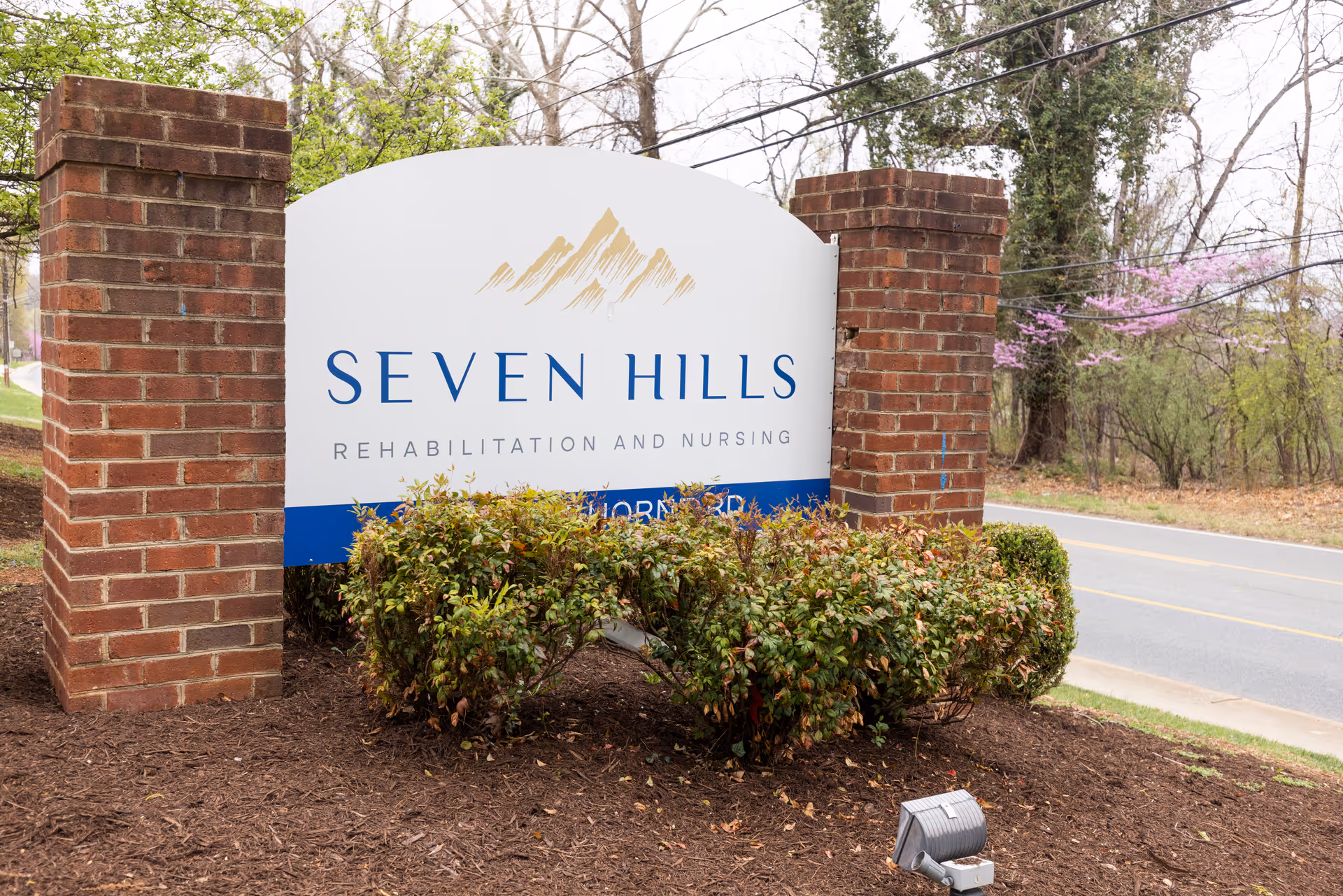 Outdoor sign for Seven Hills Rehabilitation and Nursing, mounted between two brick pillars with bushes in front and a road and trees in the background.