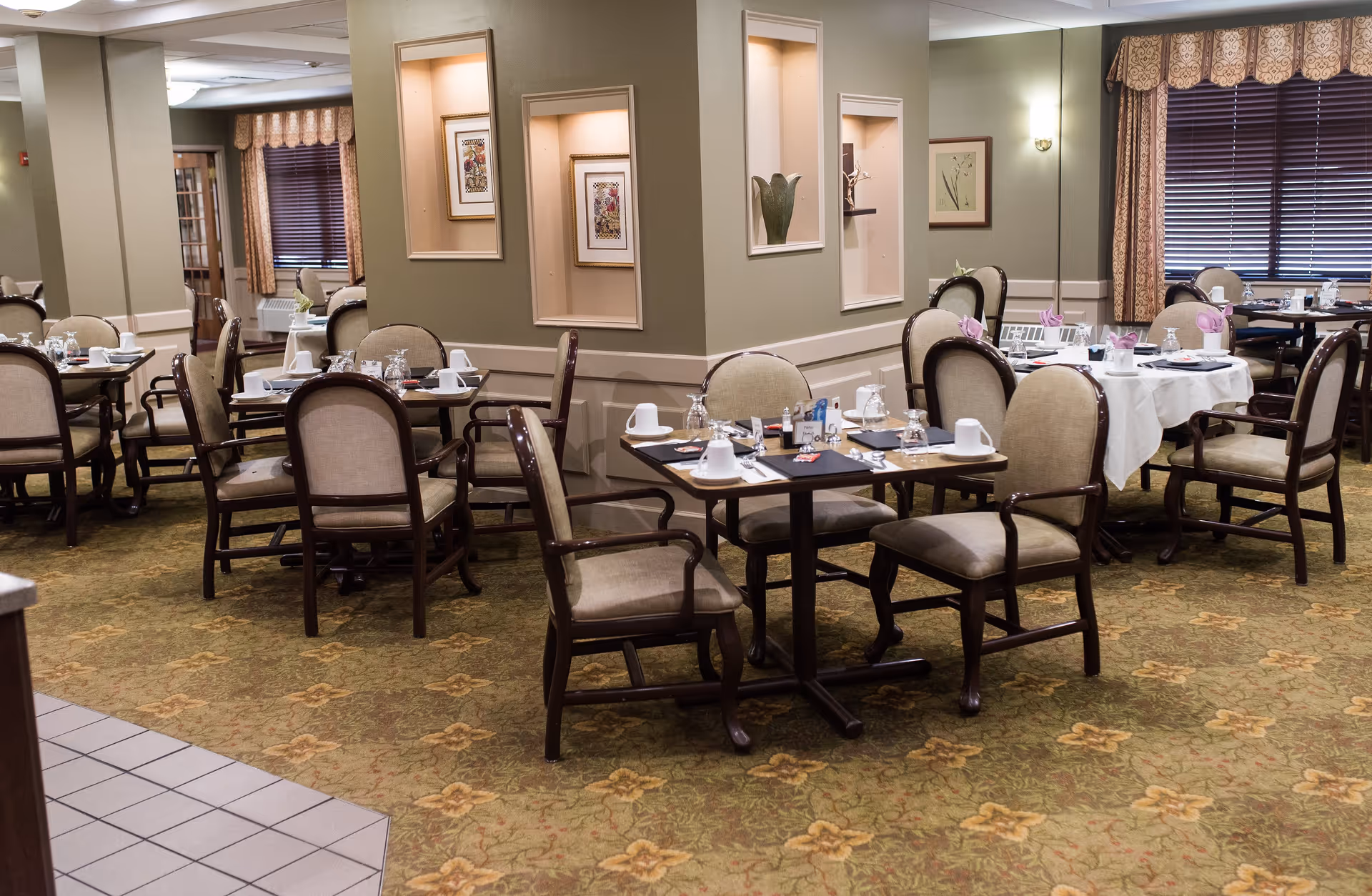 A dining room in a senior living facility with multiple tables set for meals. Each table has chairs with cushioned seats and backs, place settings including cups, glasses, and napkins. The room has patterned carpet, beige walls with decorative niches containing artwork and vases, and windows with blinds and curtains.