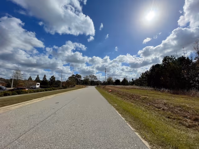 A wide paved road under a partly cloudy sky with the sun shining brightly. On the left side of the road, there are some parked cars and trees in the distance. On the right side, there is a grassy area with some bushes and trees further back.