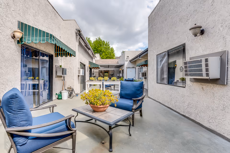 Outdoor patio area at Northridge Valley Senior Living with blue cushioned chairs, a table with a potted plant, green striped awnings over windows and doors, and air conditioning units mounted on the walls under a partly cloudy sky.