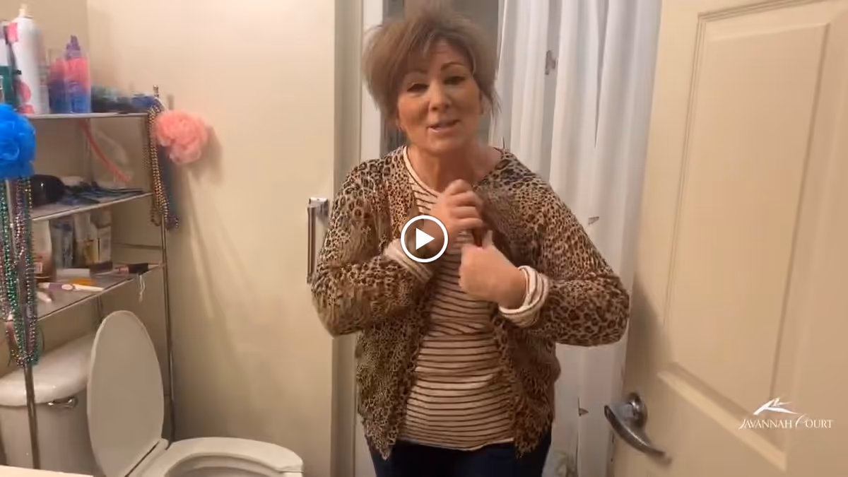 A woman standing in a bathroom next to a toilet with the lid open. There is a metal shelving unit with various toiletries and decorations, including colorful bath poufs and beads. The woman is wearing a leopard print cardigan over a striped shirt and is gesturing with her hands.